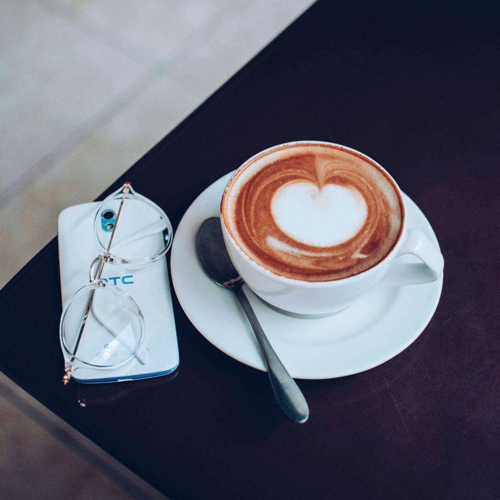 An overhead shot of a coffee cup with heart-shaped latte art next to glasses and a smartphone AND coffeehouse